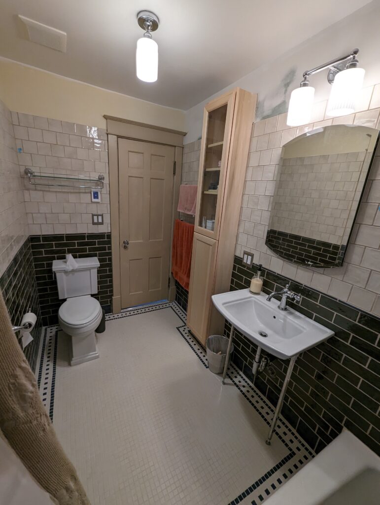 Completed vintage bathroom with a bordered mosaic floor, green tile wainscoting, new fixtures and a tall storage cabinet