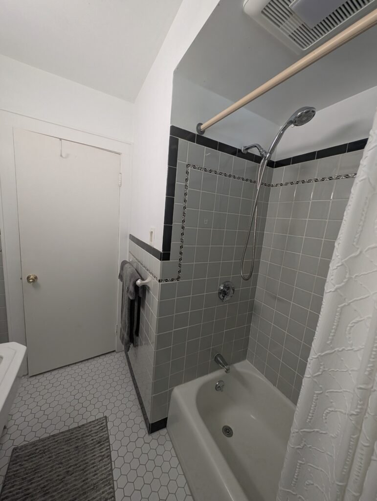 Original bathtub and shower surround with gray tile and decorative black borders before remodeling